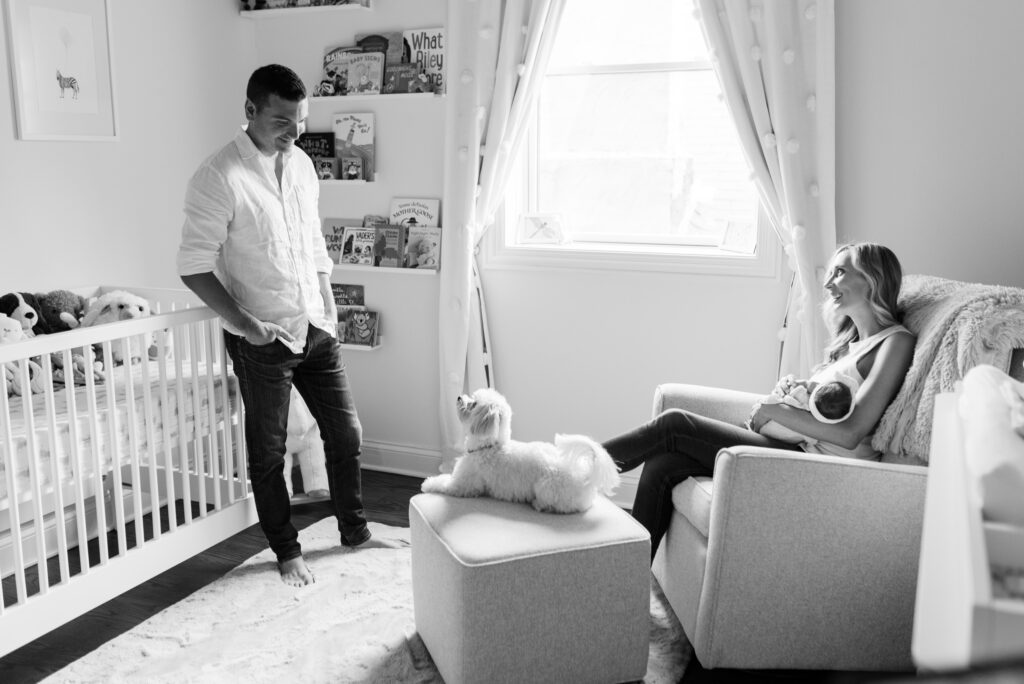 Mother holds newborn baby while she gazes at Father while he smiles at their dog.